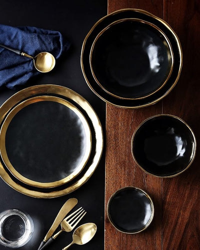 Mykonos Moonshine plates and bowls with golden finishing presented on a brown wooden and a black ground decorated with golden cutlery as well as a glass of water